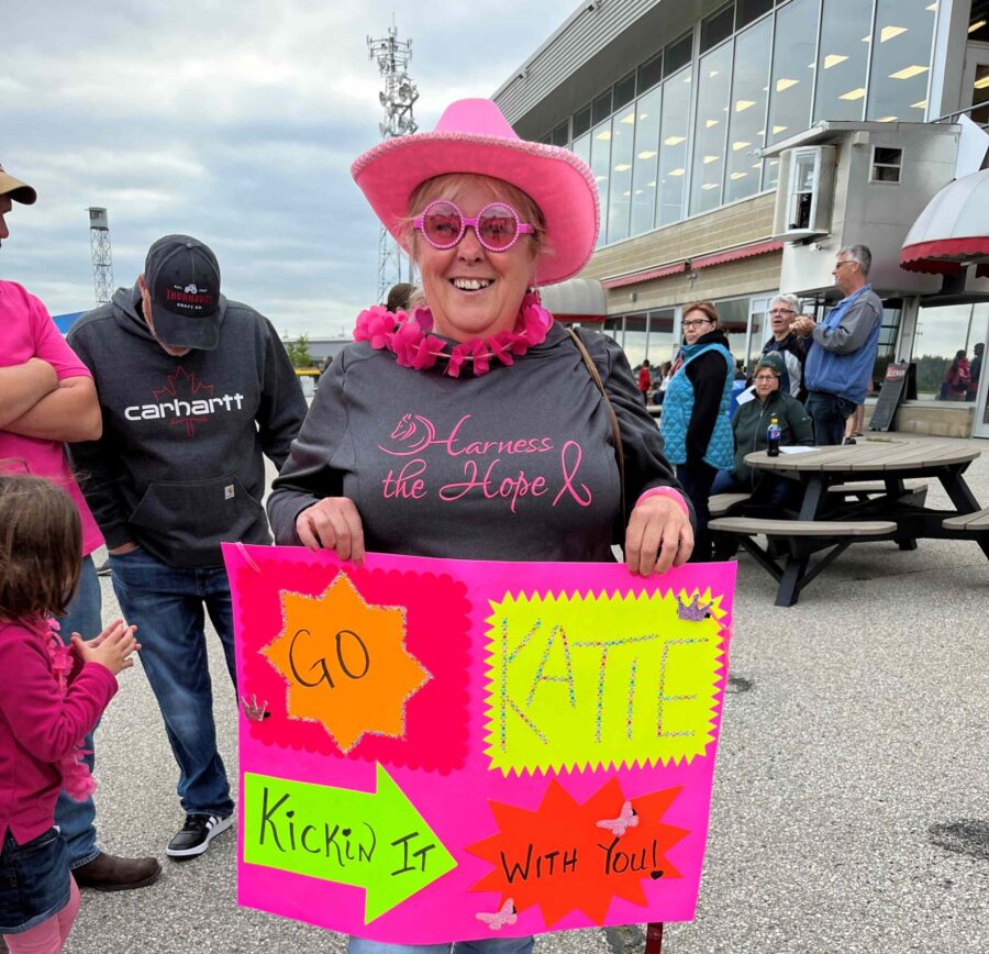 white lady with pink hat, sunglasses, grey t-shirt that says harness the hope, holding pink sign with Go katie