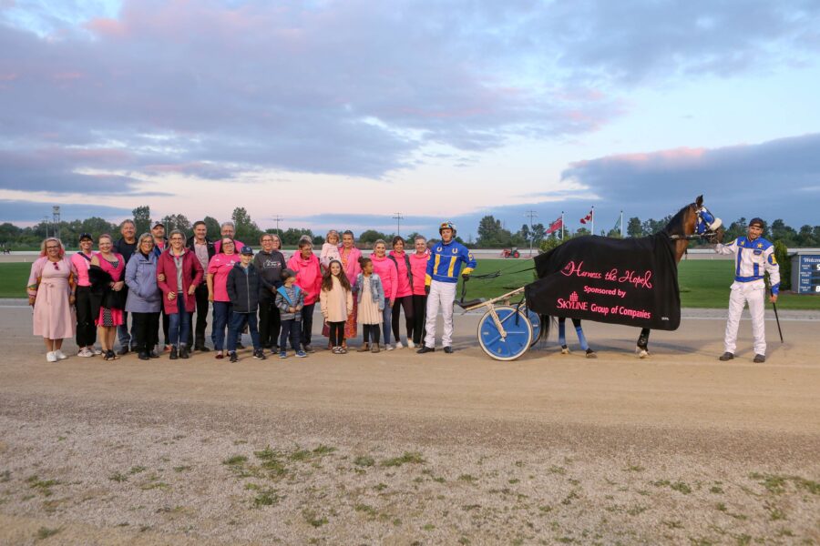 group of people in pink next to a driver and trainer horse with black blanket on it.